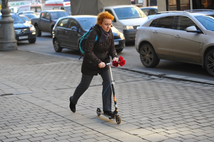 Аномально теплая зима в Москве. Женщина едет на самокате по тротуару.