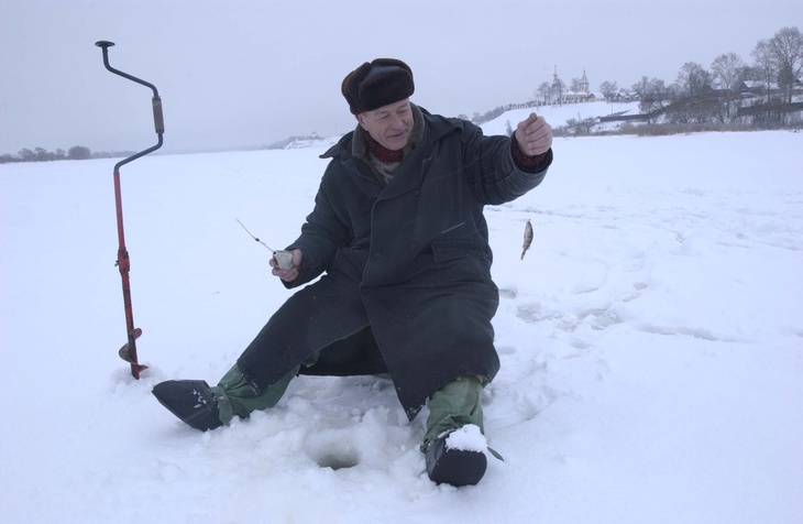 Рыбак сидит на льду с уловом в руке, рядом в лед воткнут ледоруб.
